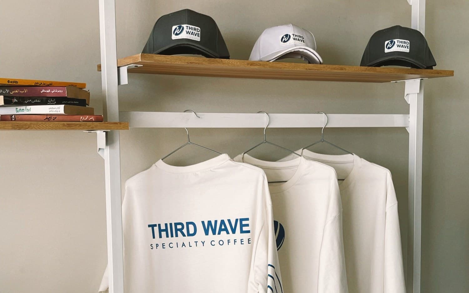 A client's branded merch display: Third Wave logo caps on a wooden shelf above white Third Wave Specialty Coffee tees on hangers